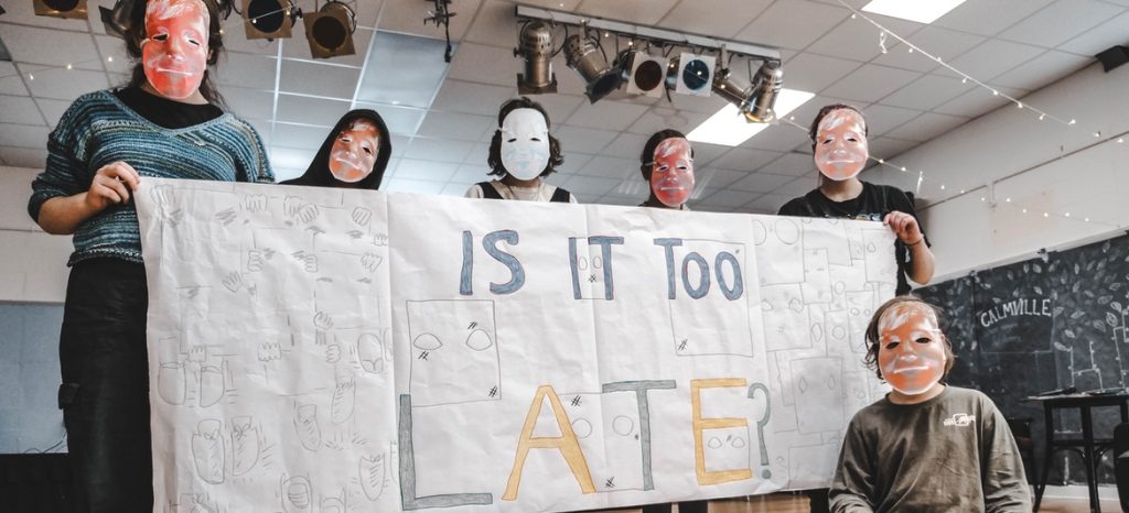 Group of young people wearing masks hold up a banner reading "Is It Too Late"