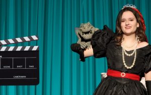 A woman in a costume holds a monster puppet as a clapperboard is raised beside her.